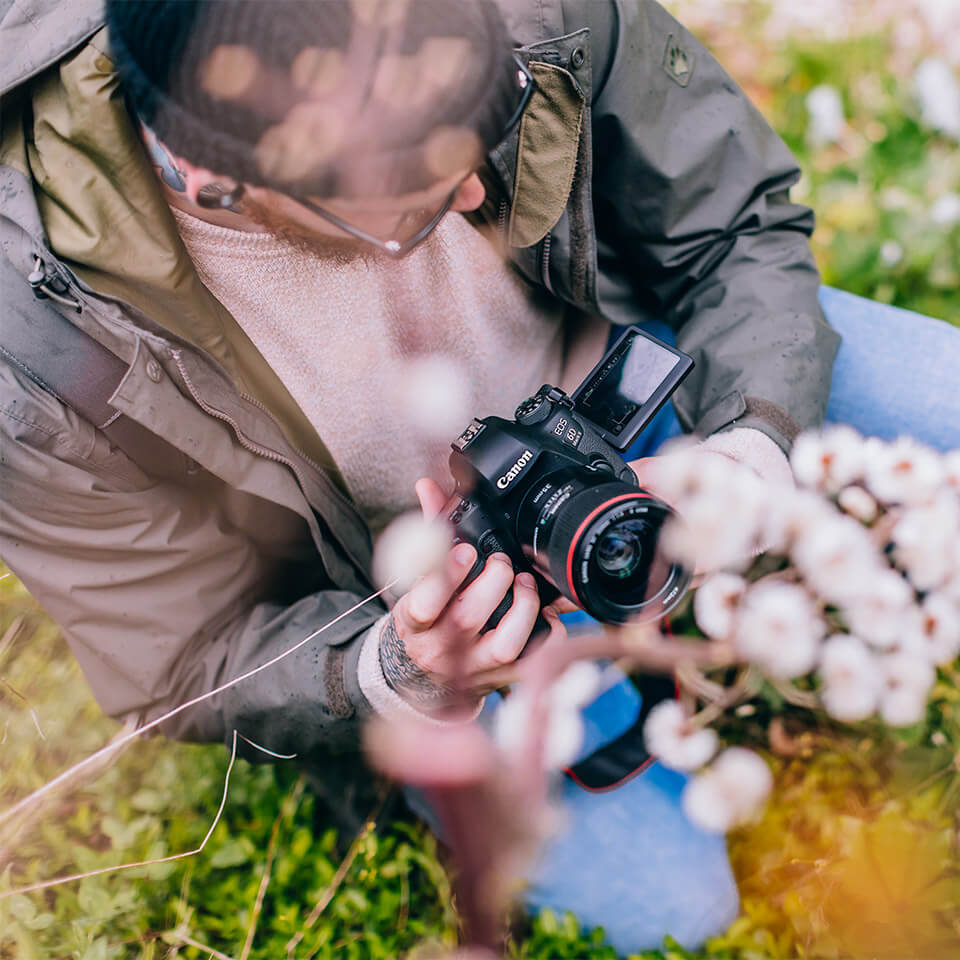 Fotograf mit Canon EOS 6D Mark II im Outdoor-Einsatz, kniet mit Kamera in der Hand vor Blüten im grünen Umfeld