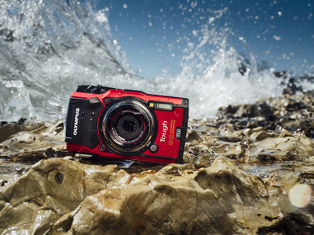 Olympus Tough TG-5 Outdoor-Kamera in Rot liegt auf nassem Felsen, während Wasser im Hintergrund spritzt