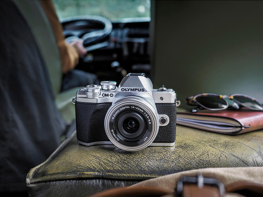 Olympus OM-D E-M10 Mark III – kompakte Systemkamera als perfekter Reisebegleiter Olympus OM-D E-M10 Mark III Systemkamera mit M.Zuiko 14–42mm Objektiv auf einem Fahrzeugsitz, daneben Sonnenbrille und Portemonnaie