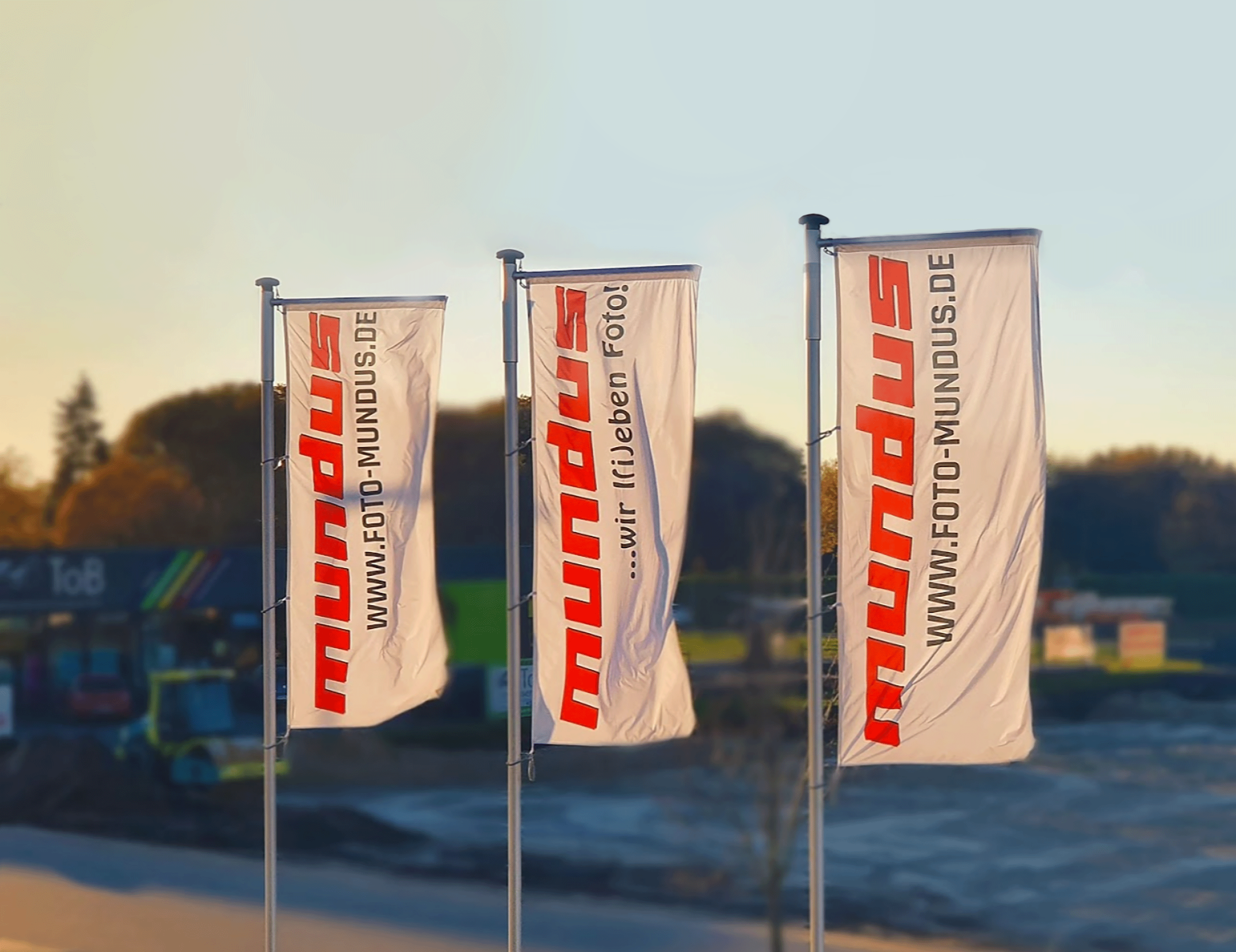 Drei wehende Foto Mundus Flaggen mit Logo und Slogan vor Sonnenuntergang – Symbolbild für neuen Onlineshop