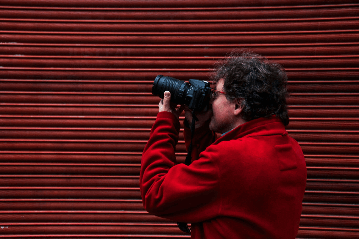 Fotograf mit Spiegelreflexkamera – idealer Einstieg in die kreative Fotografie Einsteiger-Fotograf in roter Jacke hält eine DSLR-Kamera vor eine rote Metallwand